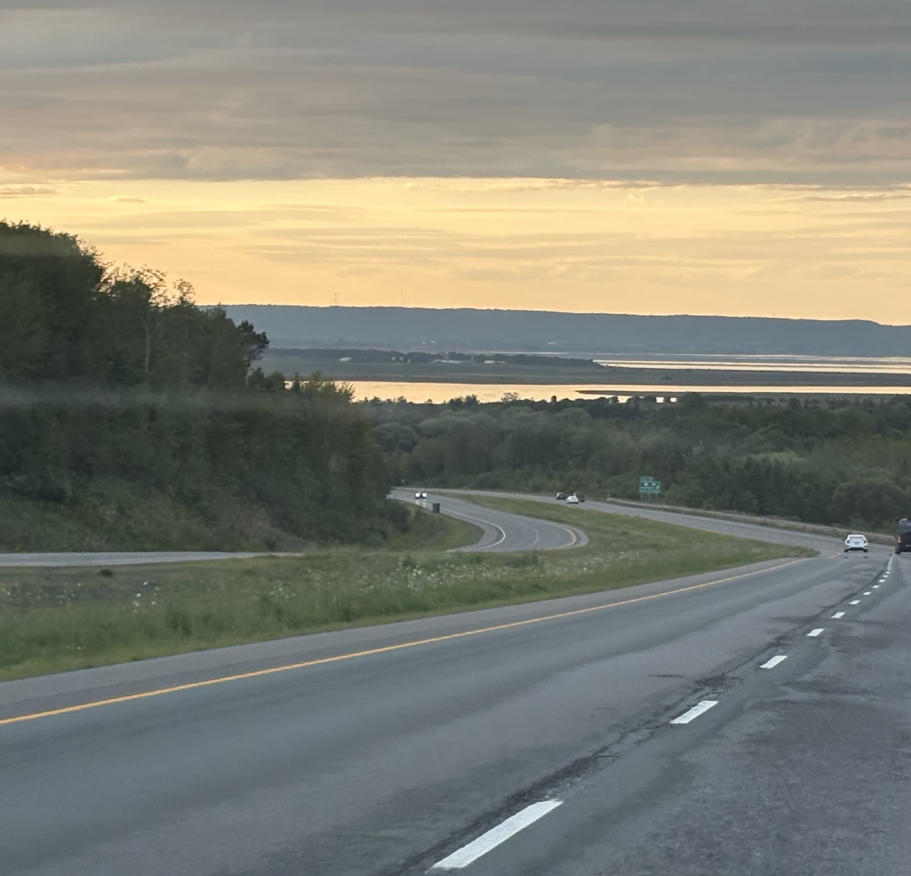 A divided highway curving downhill toward a tidal basin at dusk, Nova Scotia; a few cars far ahead