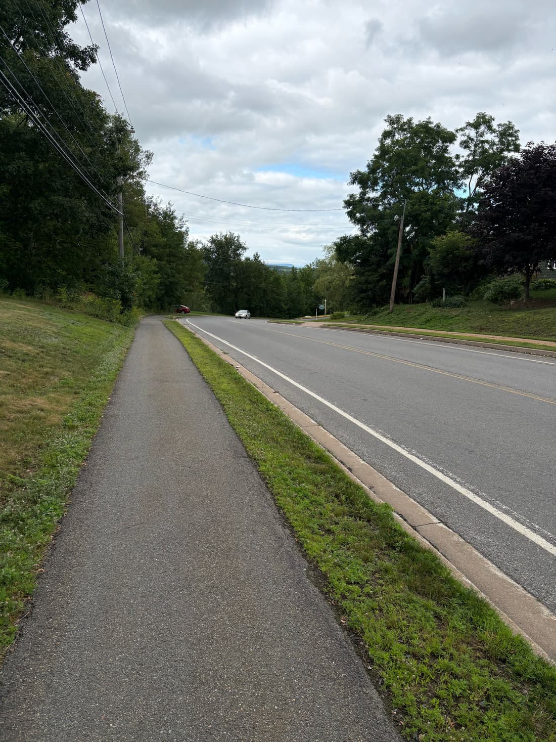 A sidewalk beside a two-lane road under an overcast sky, trees and power lines ahead